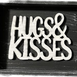May include: A black wooden tray with white distressed lettering that reads "HUGS & KISSES". The tray has a distressed white border and a black interior, creating a rustic aesthetic.