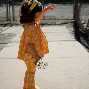 May include: A young girl wearing a yellow and pink floral print dress and matching pants with a yellow headband. She is holding a bouquet of pink flowers and is standing on a sidewalk.