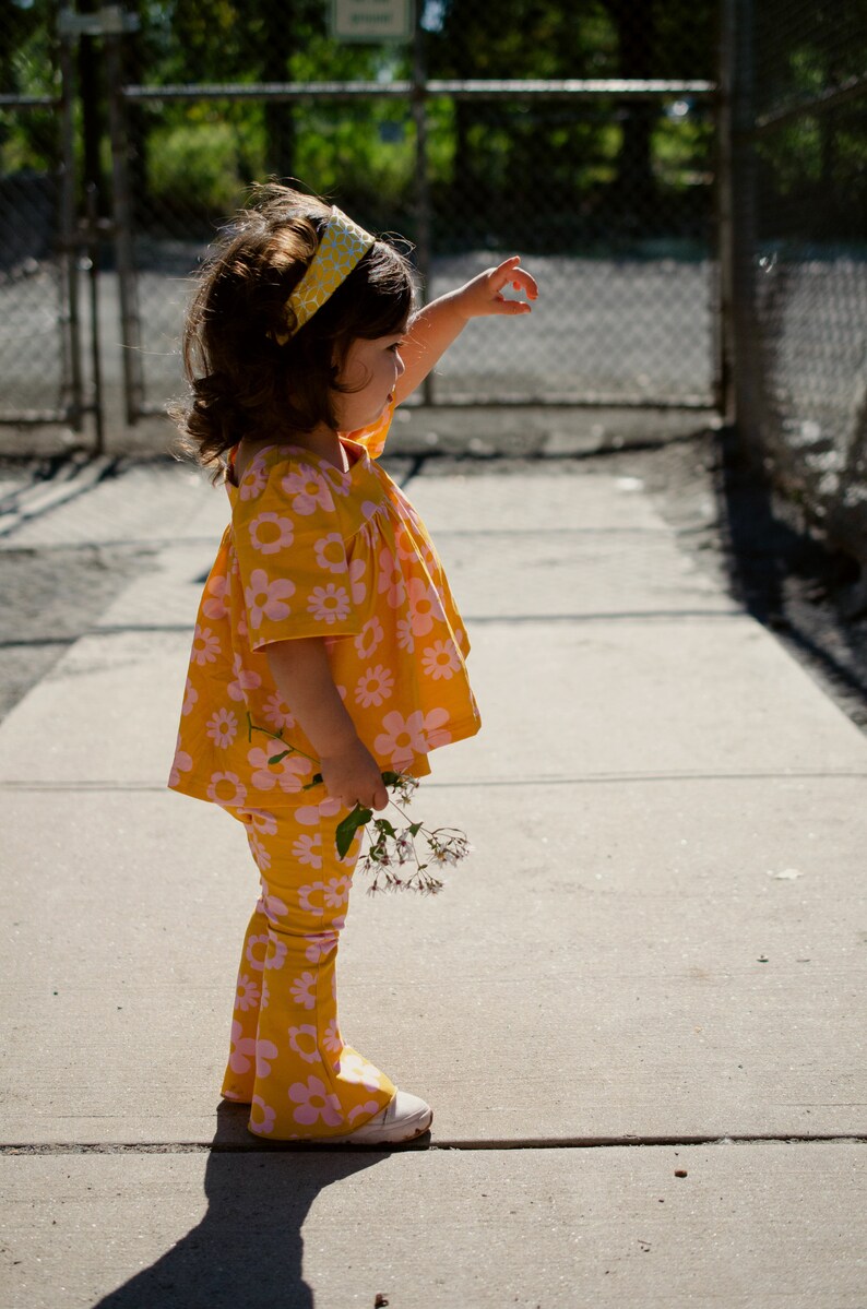 May include: A young girl wearing a yellow and pink floral print dress and matching pants with a yellow headband. She is holding a bouquet of pink flowers and is standing on a sidewalk.
