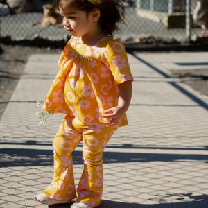 May include: A young girl wearing a yellow and pink floral print outfit with a yellow headband. She is standing on a sidewalk with a chain-link fence in the background.