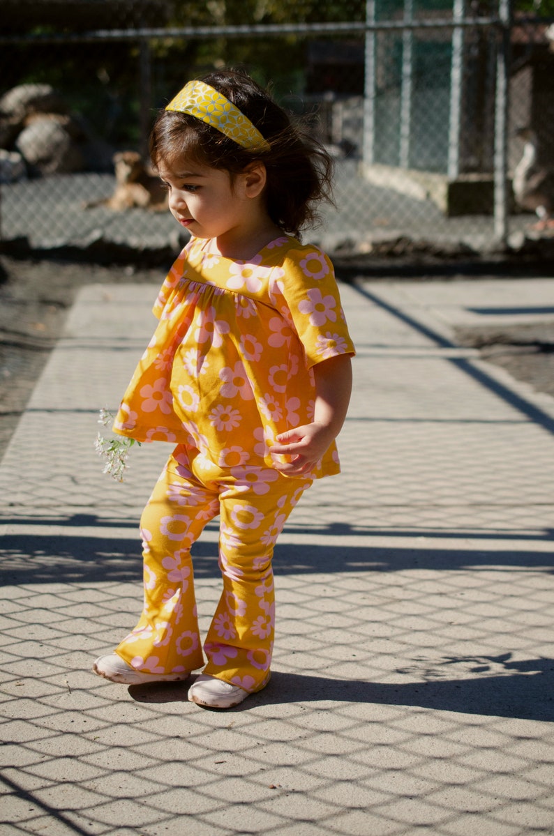 May include: A young girl wearing a yellow and pink floral print outfit with a yellow headband. She is standing on a sidewalk with a chain-link fence in the background.
