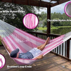 May include: A pink and white striped nylon hammock with the text "Extra Wide Nylon Bed", "Durable Nylon Arms", and "Braided Loop Ends". The hammock is suspended on a wooden deck overlooking a lake. The hammock is designed for outdoor relaxation.