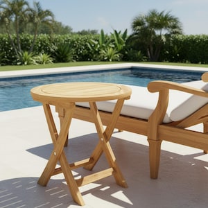 May include: A round, light-brown wooden side table with a folding X-shaped base. The table is next to a wooden lounge chair with a white cushion. The background includes a swimming pool and tropical foliage. The table is approximately 20 inches in diameter.