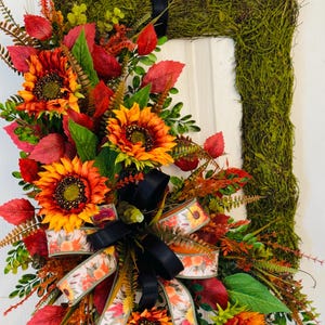 May include: A rectangular wreath base covered in green moss, decorated with artificial sunflowers in shades of orange and yellow, red and green foliage, and a multi-layered bow featuring a black ribbon and a floral patterned ribbon.