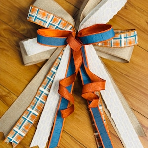 May include: A decorative bow made from multiple ribbons featuring a mix of textures and patterns. The ribbons include solid burnt orange, solid blue, white with a geometric honeycomb pattern, tan burlap, and a plaid print in orange, blue, and white.