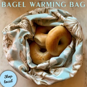 May include: A blue and brown patterned fabric bag with a drawstring closure, filled with four plain bagels. The bag is on a white marble surface. The text "Shop local" is on a blue circle in the lower left corner of the image.