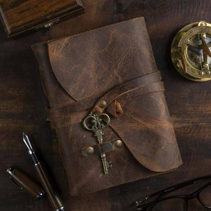 May include: A brown leather-bound journal with a key charm and a strap closure. The journal is on a wooden surface with a compass and a pen.