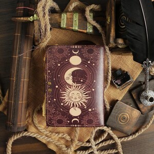 Puede incluir: Un diario de cuero burdeos con un dise&ntilde;o celestial que presenta un sol, una luna y estrellas. El diario tiene un cierre met&aacute;lico decorativo y est&aacute; rodeado de cuerda y arpillera.