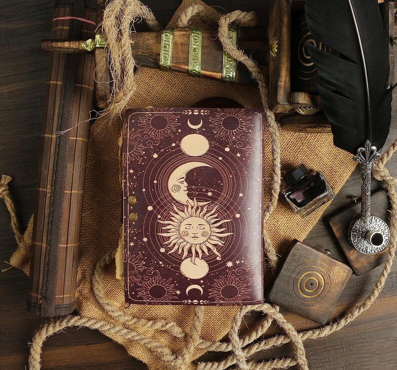 Puede incluir: Un diario de cuero burdeos con un dise&ntilde;o celestial que presenta un sol, una luna y estrellas. El diario tiene un cierre met&aacute;lico decorativo y est&aacute; rodeado de cuerda y arpillera.