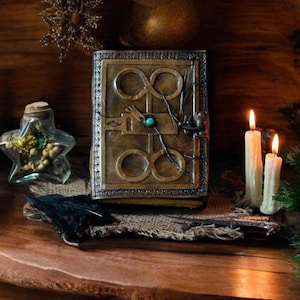 May include: A brown leather journal with a decorative metal clasp and a turquoise gemstone. The journal features an intricate design of infinity symbols and a keyhole. The journal is resting on a hessian surface with a black feather and three white candles.