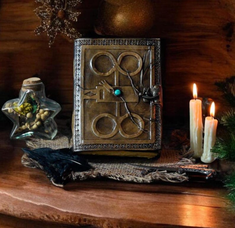 May include: A brown leather journal with a decorative metal clasp and a turquoise gemstone. The journal features an intricate design of infinity symbols and a keyhole. The journal is resting on a hessian surface with a black feather and three white candles.
