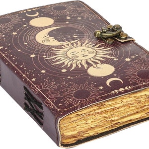 Puede incluir: Un diario de cuero burdeos con un dise&ntilde;o celestial que presenta un sol, una luna y estrellas. El diario tiene un cierre de broche y est&aacute; hecho de papel reciclado.