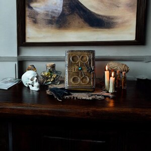 May include: A dark wooden table with a vintage book, a skull, candles, and other decorative items. The book has a leather cover with a metal design and a green gemstone.