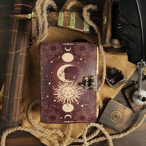 Puede incluir: Un diario de cuero burdeos con un dise&ntilde;o celestial que presenta lunas, estrellas y soles. El diario tiene un cierre de metal y est&aacute; decorado con una cuerda y detalles de madera.