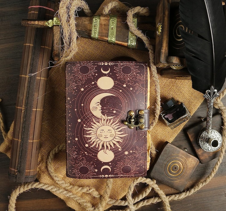 Puede incluir: Un diario de cuero burdeos con un dise&ntilde;o celestial que presenta lunas, estrellas y soles. El diario tiene un cierre de metal y est&aacute; decorado con una cuerda y detalles de madera.