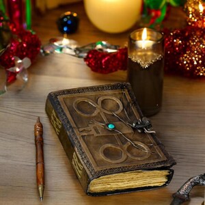 May include: A brown leather-bound journal with a decorative metal clasp and a turquoise gemstone. The journal is open to reveal blank pages. A wooden pen and a silver dagger are also in the image.