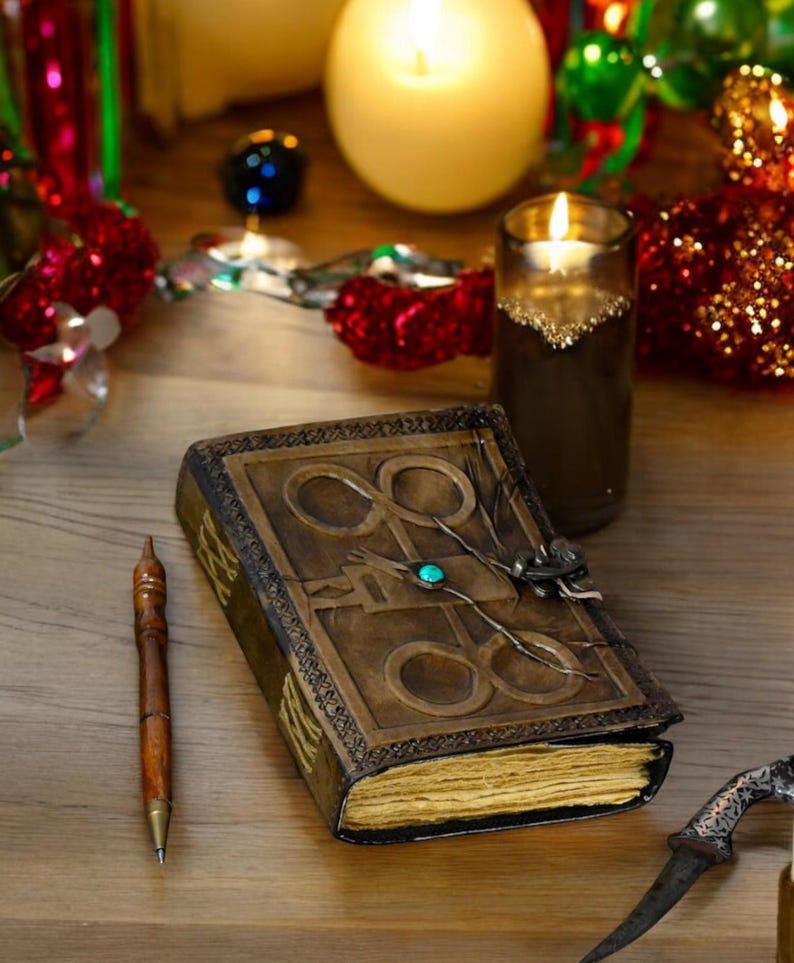 May include: A brown leather-bound journal with a decorative metal clasp and a turquoise gemstone. The journal is open to reveal blank pages. A wooden pen and a silver dagger are also in the image.