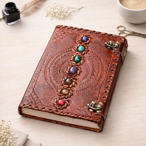 May include: A brown leather journal with a decorative embossed design and a row of colorful gemstones. The journal has a metal clasp and stitched edges. The cover features a detailed pattern with a central circular design.