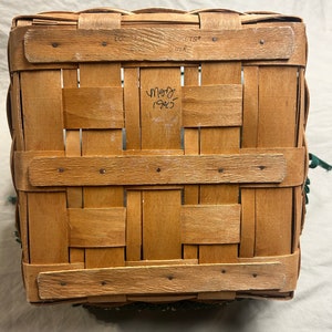 May include: A rectangular, woven wooden basket with a light brown hue. The basket features a classic weave pattern and is reinforced with horizontal wooden slats. The text "LONGABERGER BASKETS" and "MADE IN USA" is visible. The basket has a green trim.