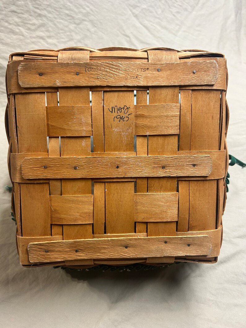 May include: A rectangular, woven wooden basket with a light brown hue. The basket features a classic weave pattern and is reinforced with horizontal wooden slats. The text "LONGABERGER BASKETS" and "MADE IN USA" is visible. The basket has a green trim.