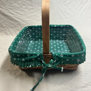 May include: A square wicker basket with a wooden handle. The basket has a green fabric liner with a white floral pattern and a ruffled edge. The liner is tied with a green fabric bow.