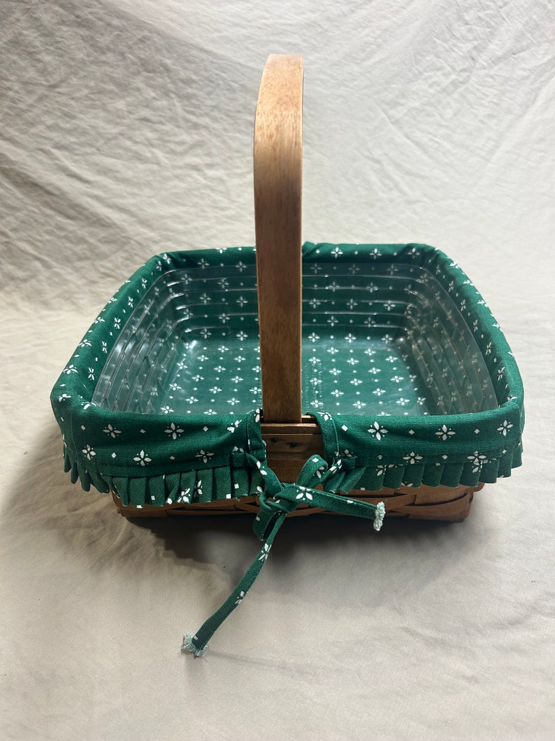 May include: A square wicker basket with a wooden handle. The basket has a green fabric liner with a white floral pattern and a ruffled edge. The liner is tied with a green fabric bow.