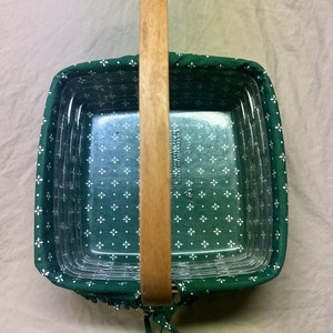 May include: Square glass baking dish with a green floral fabric cover and a wooden handle. The fabric features a white floral pattern. The handle is made of light-colored wood. The dish is designed for baking and serving.