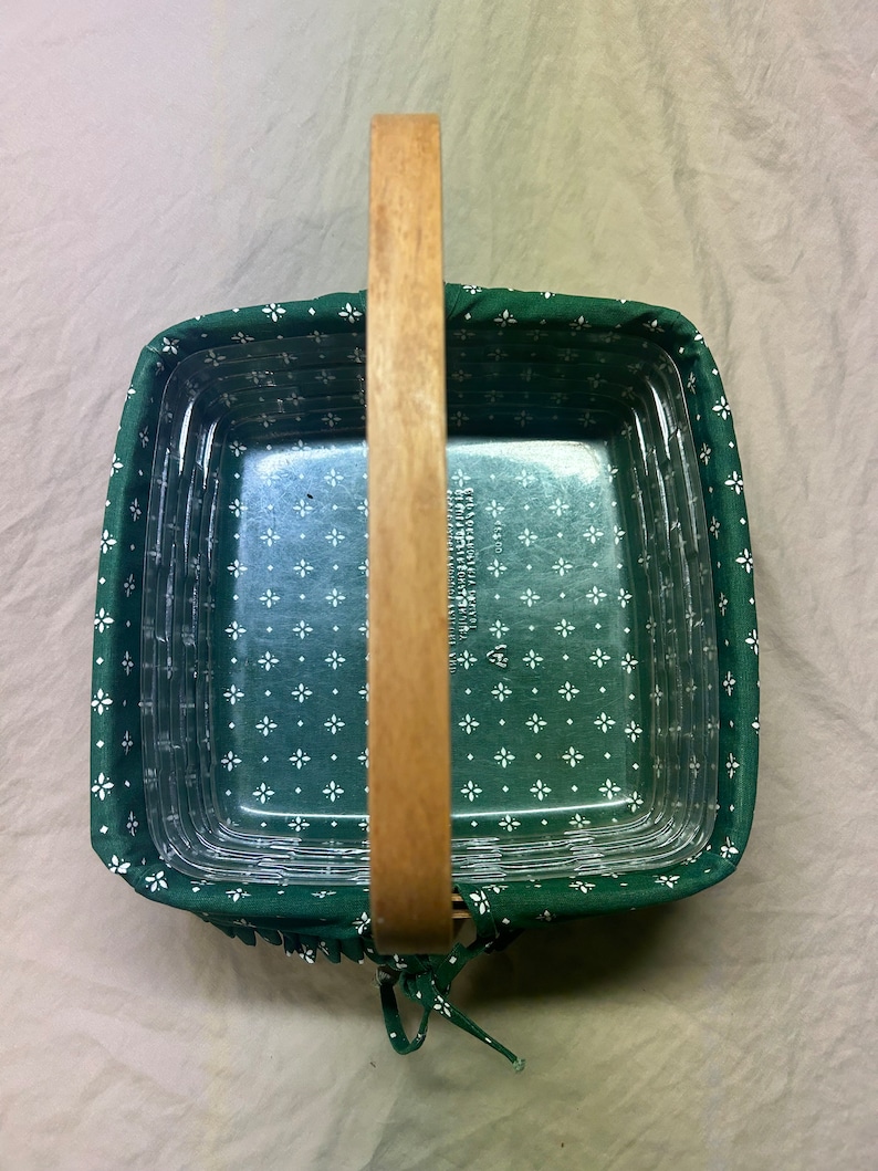 May include: Square glass baking dish with a green floral fabric cover and a wooden handle. The fabric features a white floral pattern. The handle is made of light-colored wood. The dish is designed for baking and serving.