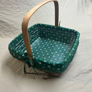 May include: Square wicker basket with a wooden handle. The interior is lined with green fabric featuring a white floral pattern. The rim is trimmed with a ruffled edge and tied with fabric bows.