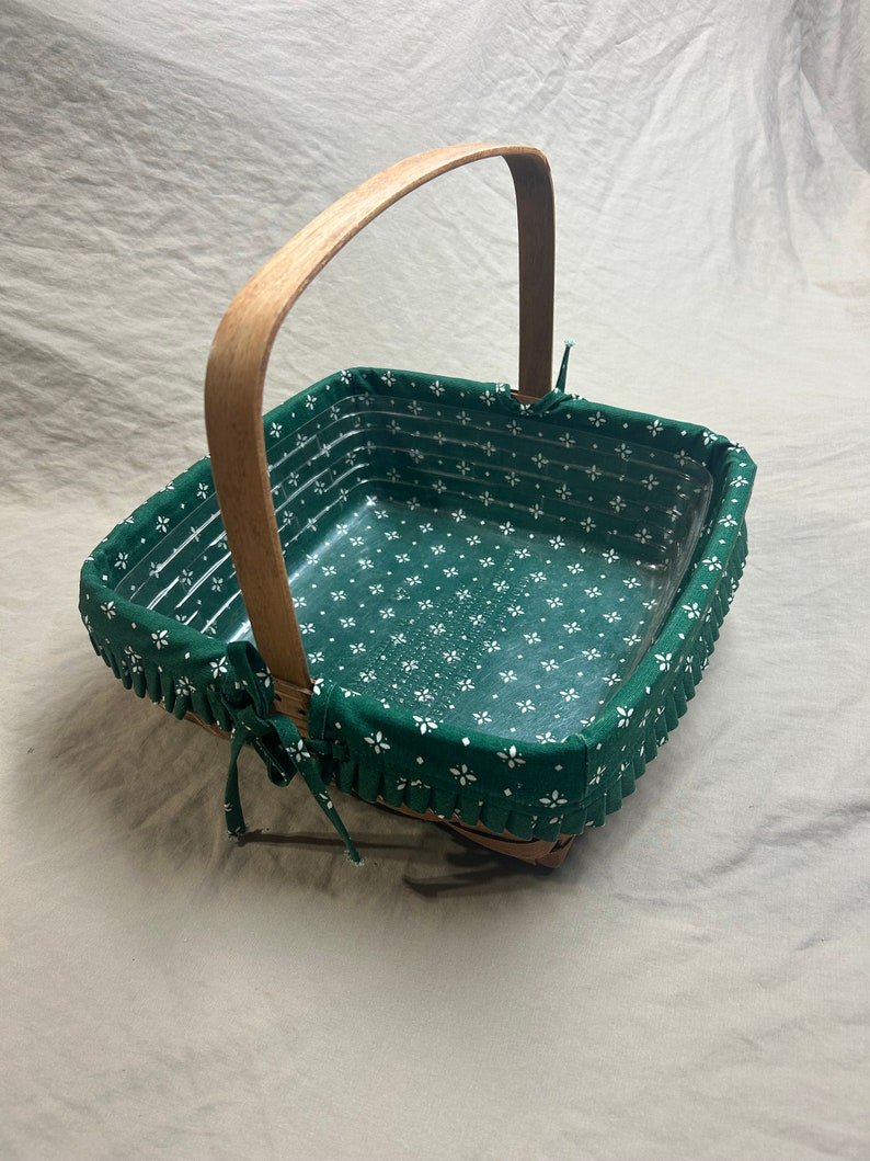 May include: Square wicker basket with a wooden handle. The interior is lined with green fabric featuring a white floral pattern. The rim is trimmed with a ruffled edge and tied with fabric bows.
