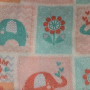 May include: A baby blanket with a repeating pattern of elephants, flowers, and the text "Dream Big little one." The elephants are teal with white accents, and the flowers are red and orange. The background is a soft pink and teal.