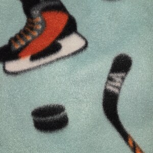May include: A light blue fleece blanket featuring a hockey theme. The design includes a hockey skate with orange and black accents, black hockey pucks, and a hockey stick with orange details.