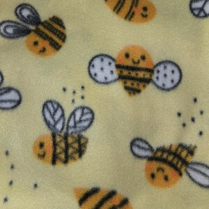 May include: Yellow fabric with a repeating pattern of cartoon bees. The bees are orange and black with white and gray wings. The background is a soft yellow color. Small black dots are scattered throughout the design.