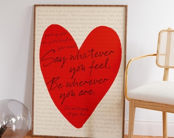 Pode incluir: Impressão emoldurada com um grande coração vermelho e o texto manuscrito "Say whatever you feel, Be wherever you are." O fundo é uma página de livro vintage. Uma cadeira moderna está à direita.