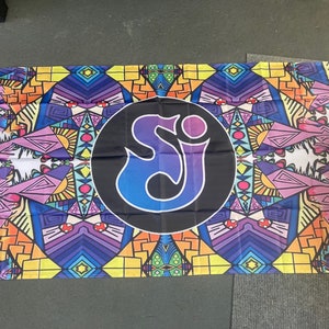 May include: A colorful flag with a geometric pattern in shades of purple, yellow, orange, and blue. The center of the flag features a black circle with a stylized "SJ" logo in blue and purple.