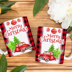 May include: Two white ceramic mugs with red and black checked designs. The mugs feature a red truck carrying a Christmas tree and the text "Merry Christmas".