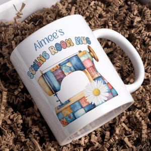 May include: White ceramic mug with the text "Aimee's Sewing Room Mug" in blue and colourful letters. The mug features a watercolour illustration of a sewing machine with a patchwork design and a daisy.