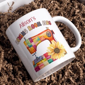 May include: White ceramic mug featuring a sewing machine design with patchwork details and a sunflower. The mug has the text "Alison's Sewing Room Mug". Presented in a box filled with brown paper.