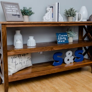 Op de afbeelding: Een houten consoletafel met twee planken. De bovenste plank heeft een wit bord met de tekst "All of me loves all of you", twee witte boekenleggers en een witte vaas met groen. De onderste plank heeft een wit kussen met zwarte tekst, twee blauwe en witte letters die "S&S" spellen, en een witte vaas. De tafel heeft een lichtbruine afwerking en een kruisvormig ontwerp aan de zijkanten.