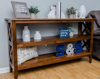 May include: A wooden console table with two shelves. The top shelf has a white sign with the text "All of me loves all of you", two white bookends, and a white vase with greenery. The bottom shelf has a white pillow with black text, two blue and white letters that spell "S&S", and a white vase. The table has a light brown finish and a cross-shaped design on the sides.