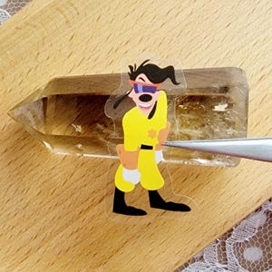 May include: A clear quartz crystal with a cartoon sticker of Goofy wearing sunglasses and a yellow jumpsuit.