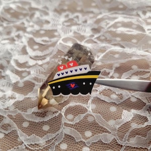 May include: A black and yellow cartoon ship sticker with red accents and a Mickey Mouse logo. The sticker is held by tweezers against a white lace background.