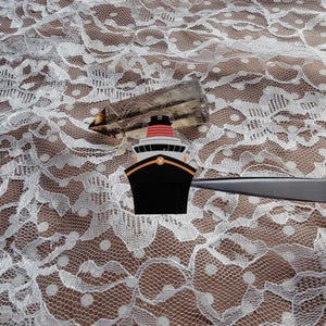 May include: A black, orange, and red cartoon ship design. The ship has a black hull, orange accents, and a red and white smokestack. The ship is set against a white lace background.