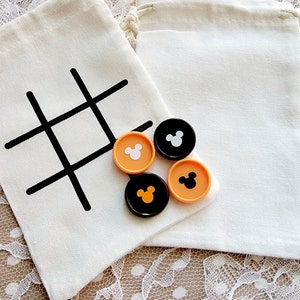 May include: Two white fabric bags with black tic-tac-toe grids printed on them. Four game pieces are on top of the bags. Two game pieces are black with white Mickey Mouse heads and two game pieces are orange with black Mickey Mouse heads.