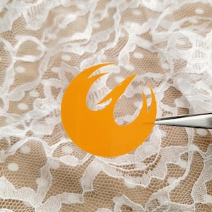 May include: A clear sticker with an orange Rebel Alliance symbol. The sticker is on a white lace background.