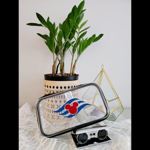 Könnte beinhalten: Eine transparente Reise-Schminktasche aus Plastik mit einem schwarzen Reißverschluss und einer Mickey-Maus-Silhouette mit einem roten Herzen auf einem blauen Wellendesign. Die Tasche liegt auf einer weißen Spitzenoberfläche mit einem schwarzen Fernglas.