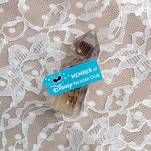 May include: A clear, faceted crystal with a light brown hue, accompanied by a turquoise tag. The tag reads "MEMBER of Disney Vacation Club" with the Disney logo. The crystal is set against a white lace background.