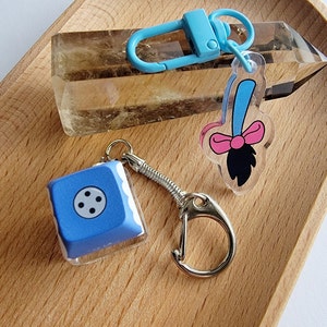 May include: A blue keycap with four black dots on a clear acrylic keychain. The keychain is attached to a silver spring ring and a silver lobster clasp. A blue and pink cartoon broom charm is attached to a separate blue keychain.