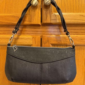 Pode incluir: Uma bolsa preta com superfície texturizada e alça trançada. A bolsa apresenta uma aba superior curva, um fecho de zíper prateado e um pequeno pingente de metal em forma de coração preso ao puxador. A alça é fixada com clipes de metal prateados.