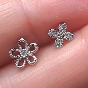 Könnte beinhalten: Ein Paar silberne Ohrringe, einer in Form einer Blume mit einem kleinen, klaren Stein in der Mitte und der andere in Form eines vierblättrigen Kleeblatts mit klaren Steinen an jeder Spitze.